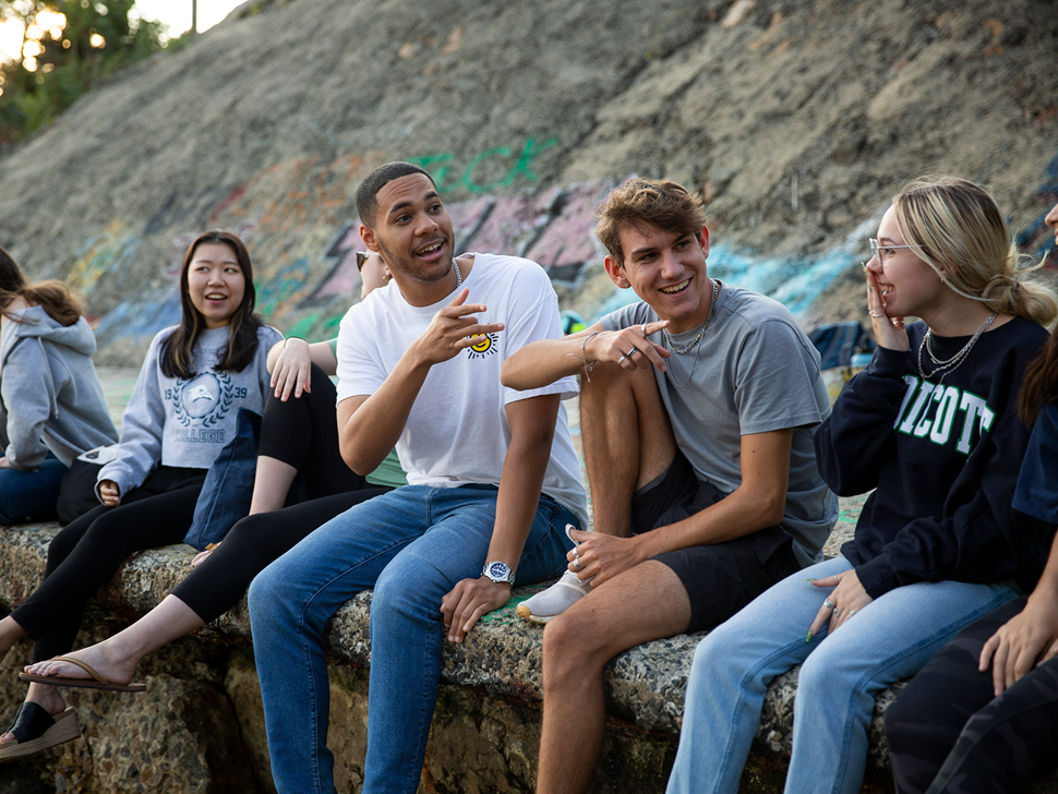 A diverse group of college students sit together outdoors, smiling and chatting.