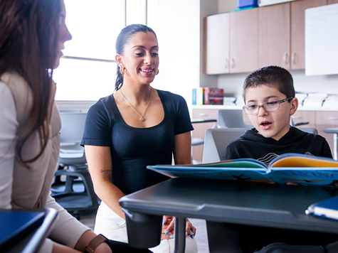 A young boy with glasses reads a large open book aloud at a desk, while a smiling teacher and another student listen attentively.