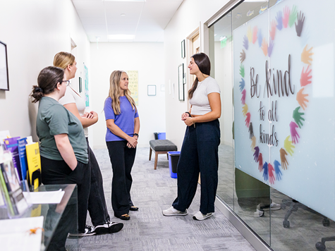 Four women stand chatting in a hallway next to a glass wall featuring a handprint heart decal that reads "Be kind to all kinds."