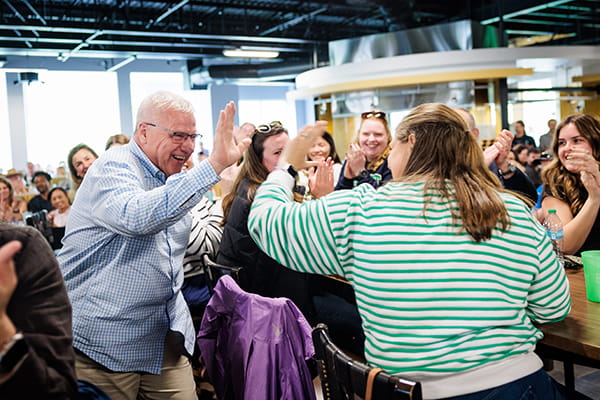 A smiling man in a light blue checkered shirt gives a high five to a woman in a green and white striped sweater in a crowded room.