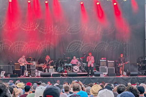 A band performs on an outdoor stage illuminated by red spotlights, viewed from the audience.