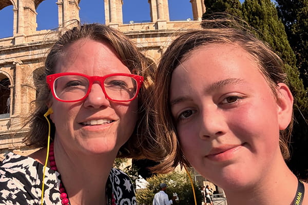 Selfie of a woman wearing red glasses and a child standing in front of the Roman Colosseum on a sunny day.