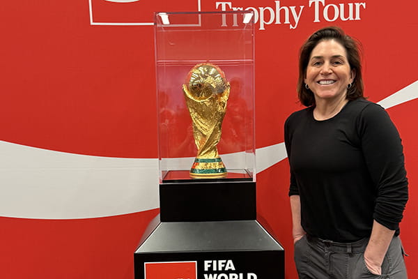 A woman stands smiling next to the FIFA World Cup trophy displayed in a glass case.
