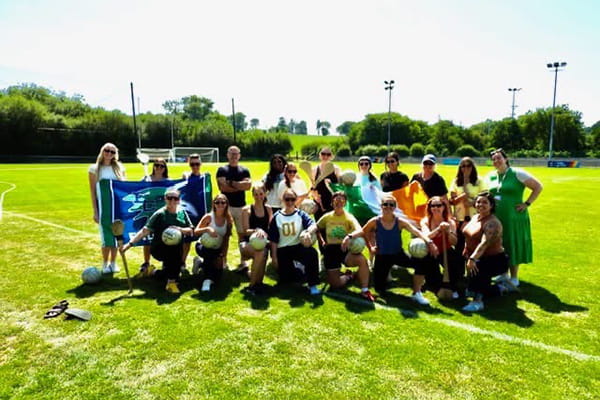 A group poses on a sunny sports field holding Gaelic footballs, hurley sticks, and Irish flags.