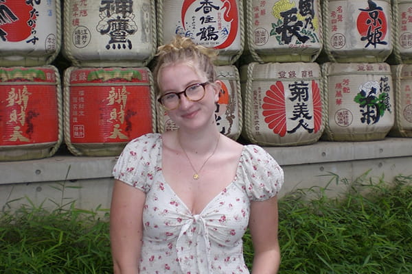 A smiling young woman with glasses and a floral top in front of a sake barrel wall.