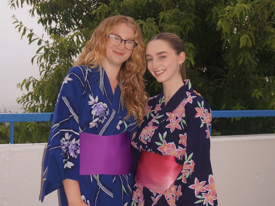 Two young women smiling in traditional Japanese kimonos with colorful obi belts against green leaves.
