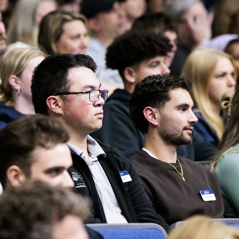 A diverse group of students and attendees sit in an audience, listening intently to a speaker.