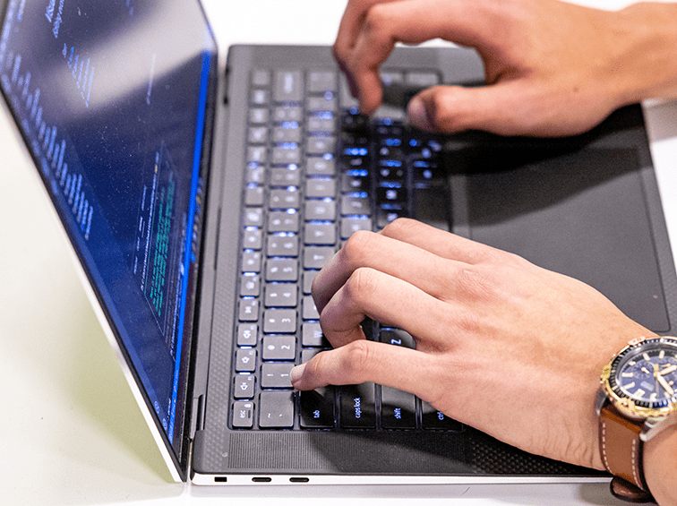 A person's hands are typing on a backlit laptop keyboard, with code visible on the screen.