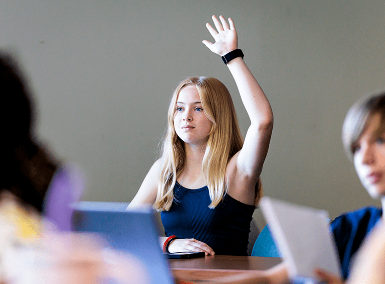 A young girl with long blonde hair and blue eyes sits at a desk, raising her hand during a workshop. She wears a black tank top and looks attentively toward the front.