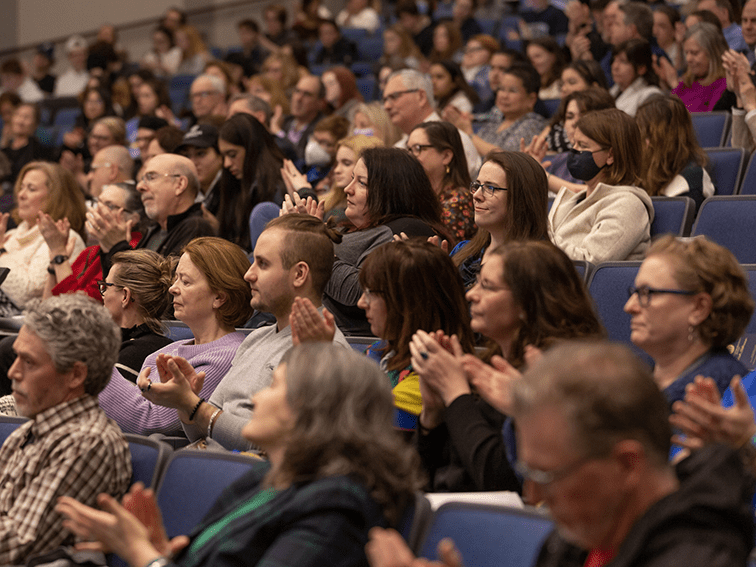 A seated audience of diverse people, some wearing glasses, are clapping in a large auditorium.