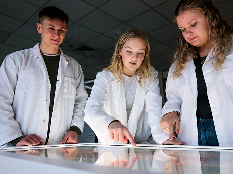 Three students in white lab coats gathered around an interactive touch table in a science lab.
