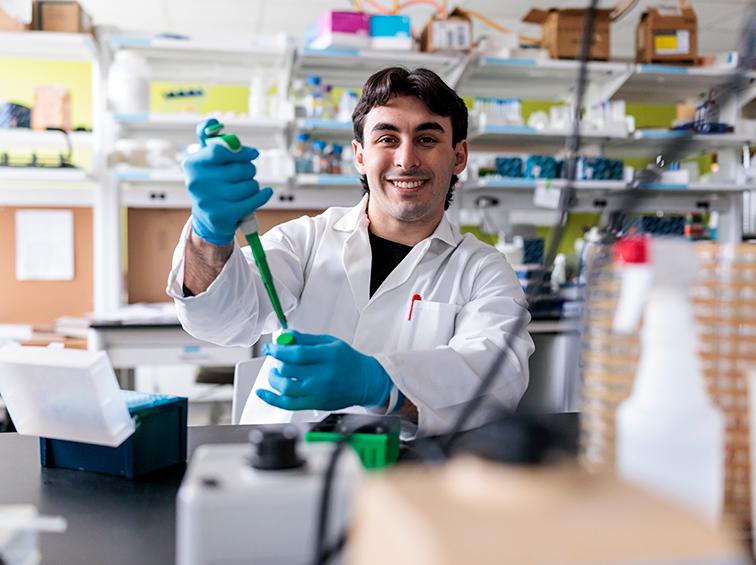 A smiling male student in a lab coat and blue gloves using a micropipette at a lab bench.