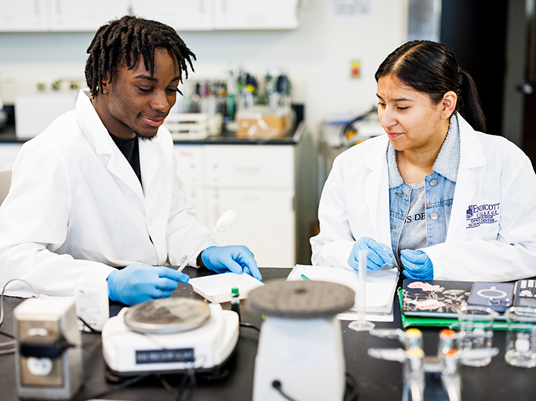 Two science students in lab coats and gloves conducting an experiment at a lab workstation.