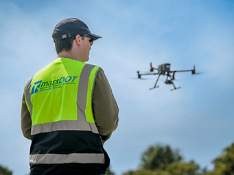 A MassDOT Aeronautics Division worker in a neon safety vest watches a drone flight against a blue sky, demonstrating aerial infrastructure inspection technology.