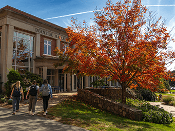 Three students with backpacks walk past a building in autumn, with a tree displaying red leaves.
