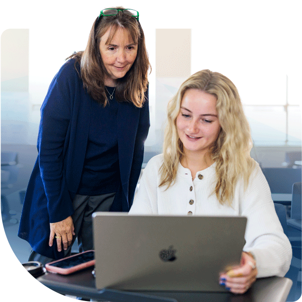 A woman with green glasses and a younger woman with blonde hair laugh together while looking at a laptop screen in a bright, indoor classroom or office setting.