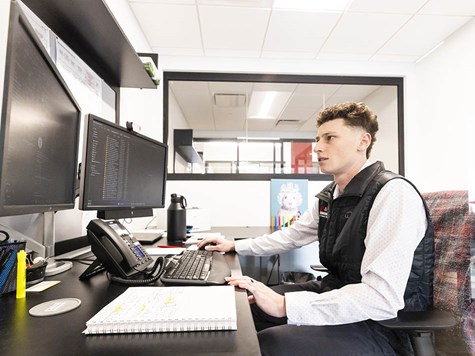 A young man wearing a vest seated at an office desk, working on dual computer monitors with a notepad nearby.