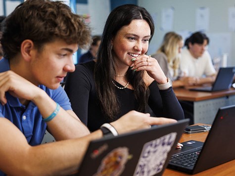 A young woman smiling at a laptop screen alongside a male classmate pointing at the display.