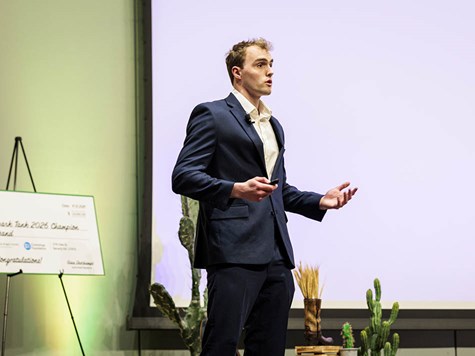 A young man in a dark blue suit speaking on stage during a business pitch presentation.