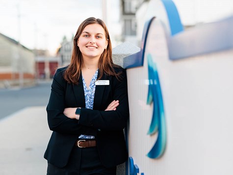 A young woman in a professional blazer and name tag smiling with arms crossed next to a blue branded sign.