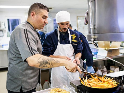 Two chefs working together in a commercial kitchen, using tongs to cook sliced vegetables in a pan.