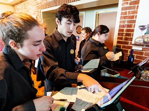 Restaurant staff members in dark uniforms working together behind a counter using a digital touchscreen terminal.