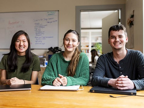 Three young adults sitting at a wooden table with laptops, smiling together toward the camera.