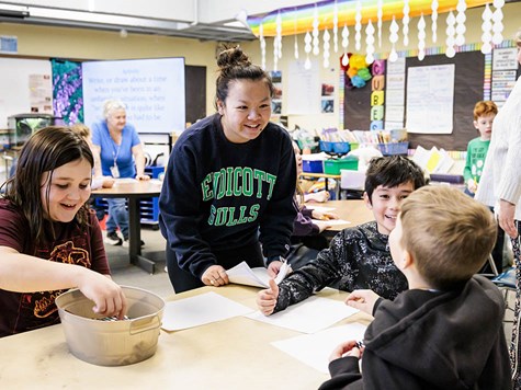 A smiling teacher in an Endicott sweatshirt leaning over a table to assist young elementary students in a classroom.
