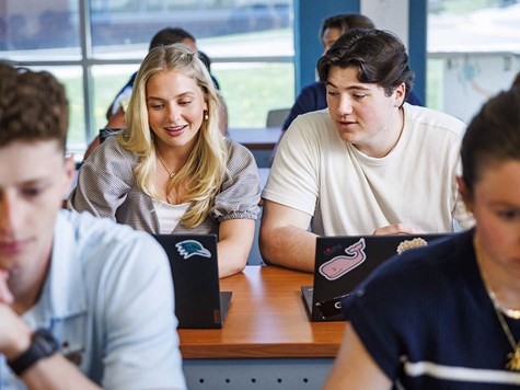 A blonde female student and a male student sitting side-by-side at a desk, looking at a shared laptop screen in class.