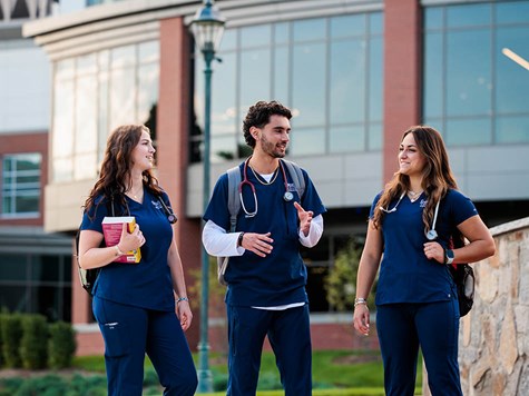 Three nursing students wearing blue scrubs walk outside together while chatting and holding books.