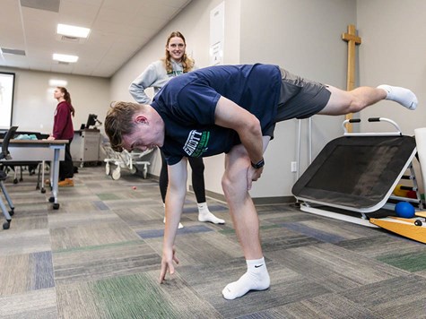 A young man balances on one leg reaching for the floor while a female instructor observes in a clinic setup.