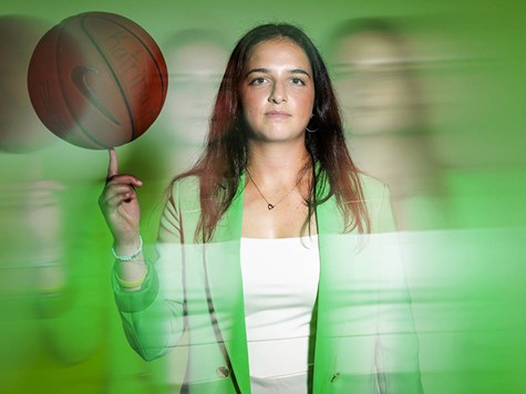 A woman in a green blazer looking forward while spinning a basketball on her index finger.
