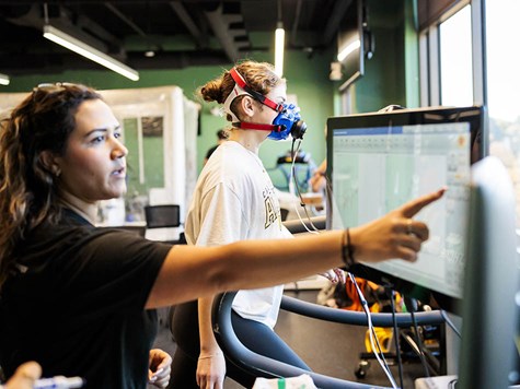 A woman wearing an oxygen mask on a treadmill while an instructor points at a screen tracking metrics.