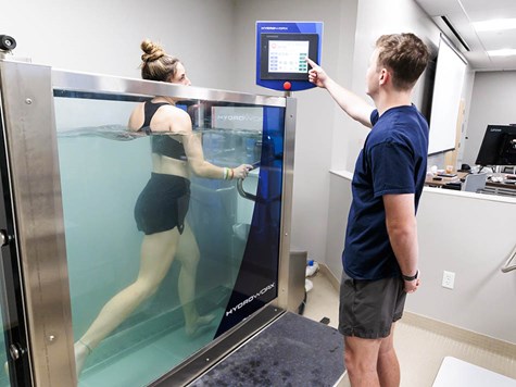 A woman walking on an underwater treadmill inside a glass tank as a man operates the control panel.