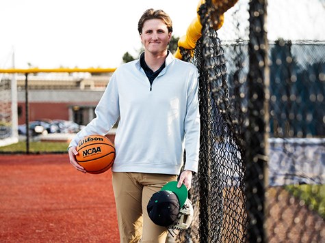 A young man smiling next to a fence while holding a basketball, baseball cap, and glove.