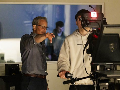 An instructor pointing in a media studio while a student operates a large broadcast camera.