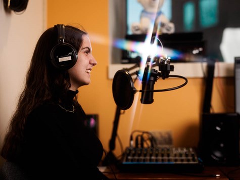 A smiling female student wearing headphones seated at a podcasting setup with a microphone and mixer.