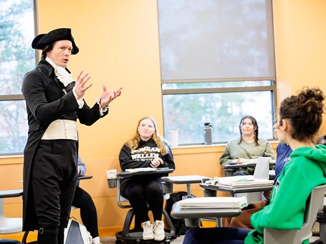 A man dressed in historical 18th-century attire lecturing to a room of college students.