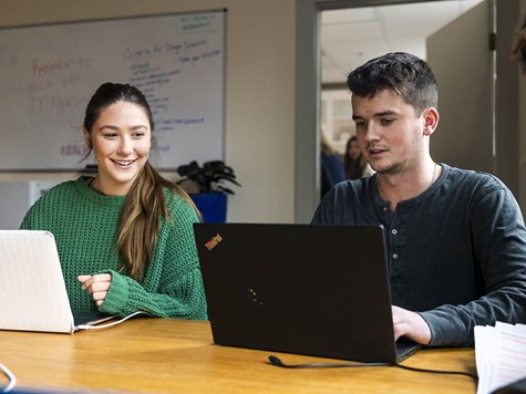 A female and male student sitting side-by-side at a table collaborating on their laptops.