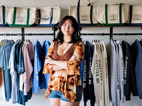 A young woman standing with arms crossed in front of clothing racks inside a retail shop.
