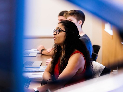 A female student with glasses sitting in a classroom, actively speaking during a discussion.