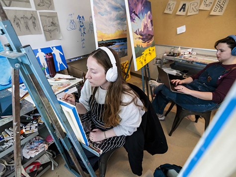 A female art student wearing white headphones sitting at an easel and painting on a canvas.