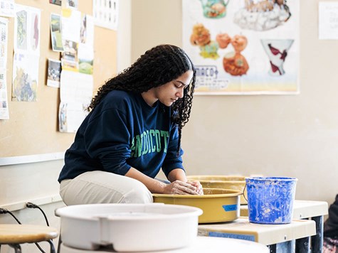 A female student leaning over a pottery wheel shaping clay in an art studio.