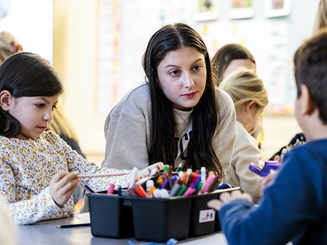 A female student leaning over a table in a classroom to assist young children with art supplies.