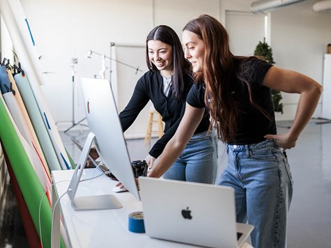 Two women standing together at a desk reviewing designs on a desktop computer monitor.