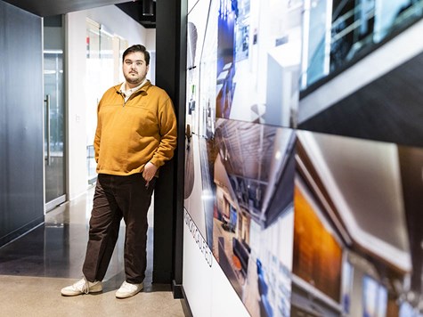 A young man leaning against a wall displaying photos of interior design projects.