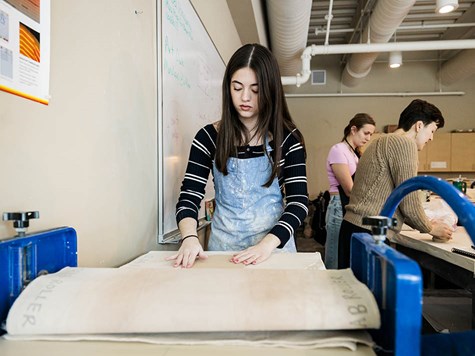 A female student using a large blue slab roller press to flatten clay in an art studio.