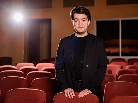 A young man in a dark blazer leaning against red seats in an empty auditorium.