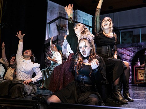 Cast members in dark Victorian-style costumes singing dramatically on stage.