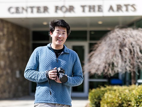 A smiling male student holding a DSLR camera outside a building labeled Center for the Arts.